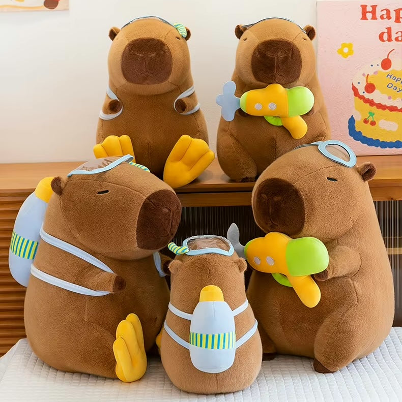 Four brown capybara plush toys arranged together on a bed, each wearing a light blue swim cap and goggles and carrying a yellow and green toy water gun or backpack accessory.