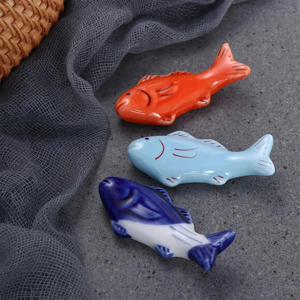 Three ceramic fish kawaii chopstick holders in orange, blue, and purple on a textured surface.