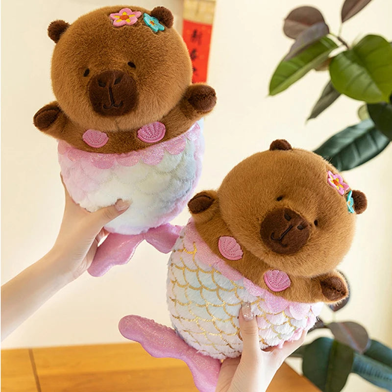 Two capybara plush toys resembling bears with mermaid tails held by a hand against a blurred indoor background.