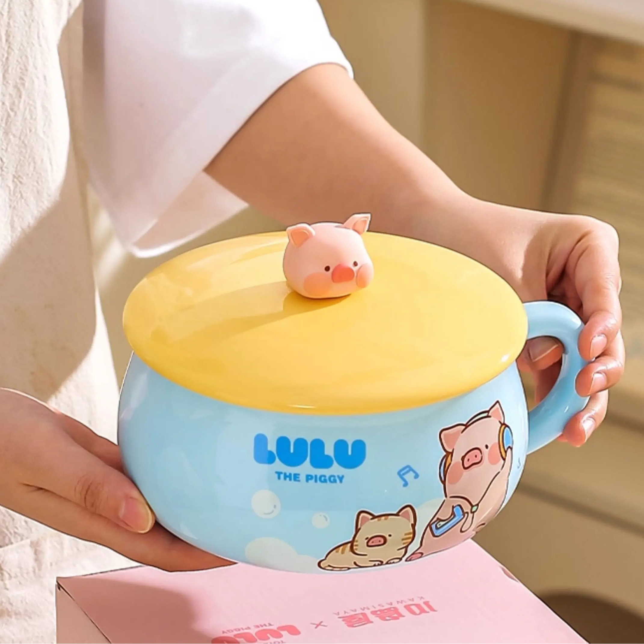Person holding a blue kawaii ramen bowl with a yellow lid featuring cartoon characters, including a pig.