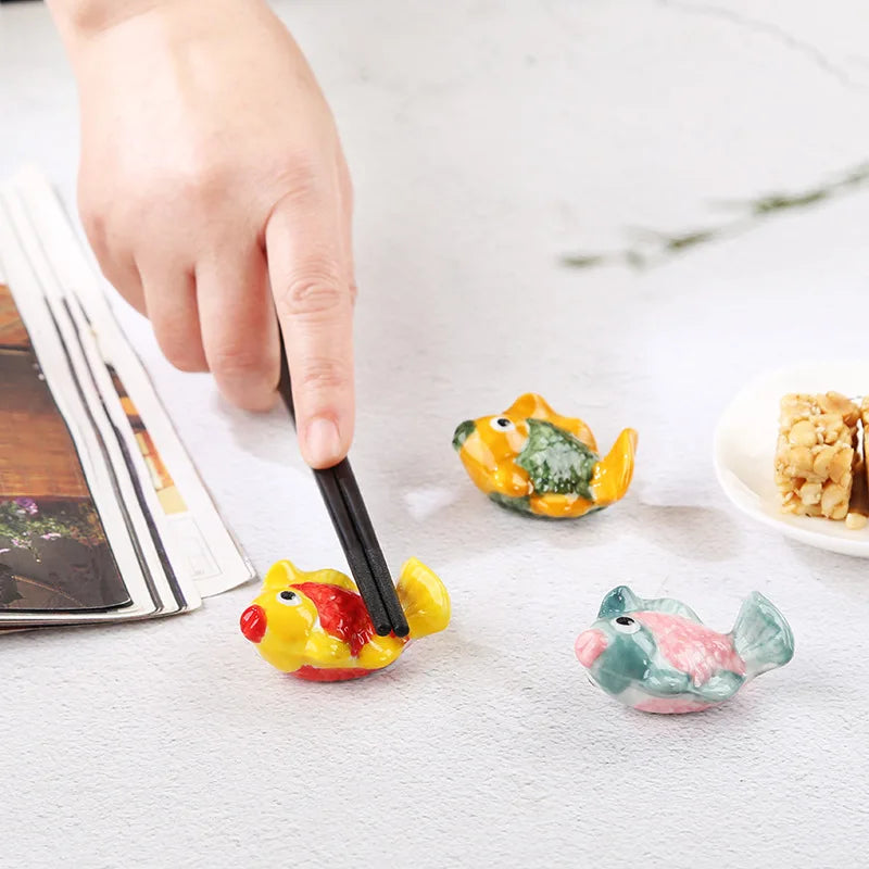 Colorful fish-shaped kawaii chopstick holders on a table with a hand holding a black tool.