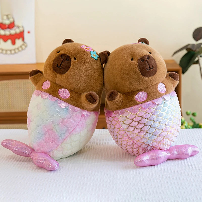 Two brown capybara plush toys with mermaid tails on a white surface.