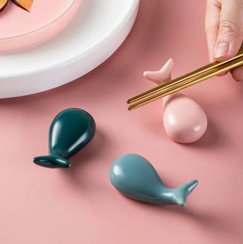 Two green whale-shaped ceramic kawaii chopstick rests on a pink surface with a hand holding gold chopsticks.