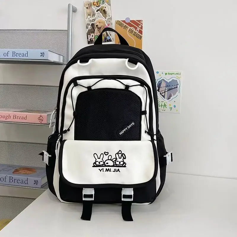 A pastel black and white school backpack with pink zipper accents and cartoon bunny graphics on the front pocket, displayed on a white table against a white wall with decorative books and cards.