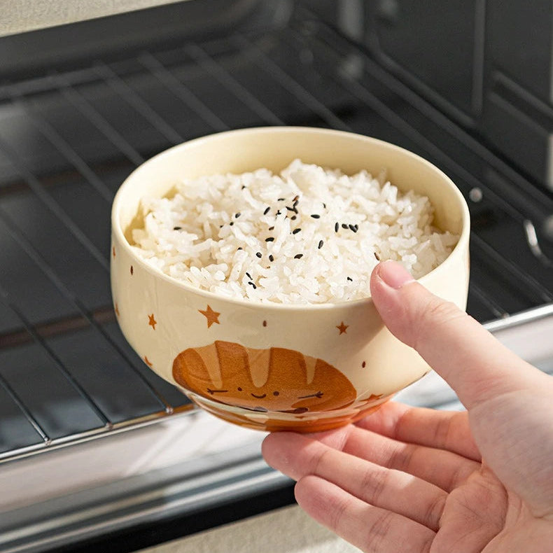 Hand holding a kawaii bowls of rice with a cartoon design in front of an oven.