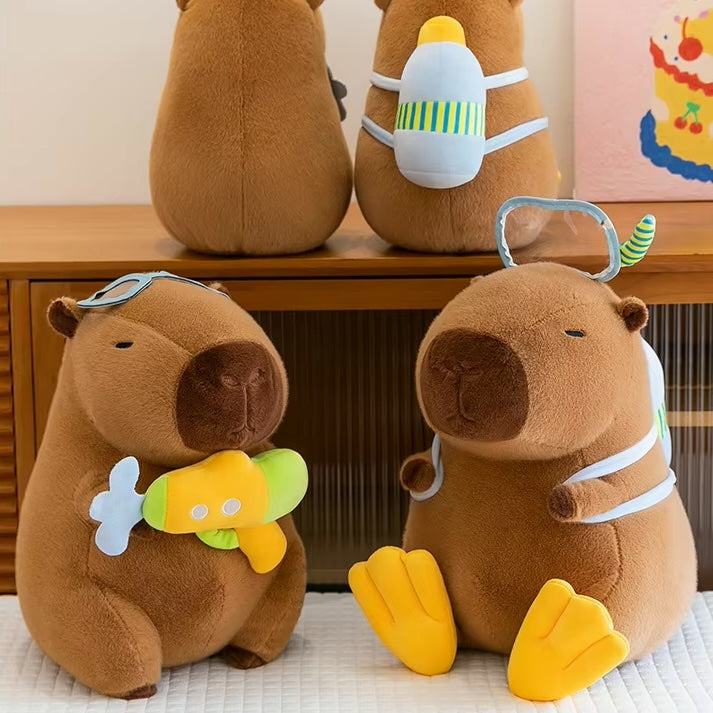 Three brown capybara plush toys with swim-themed accessories, including goggles, a snorkel mask, and a small yellow-and-green toy, arranged on a wooden shelf and surface.