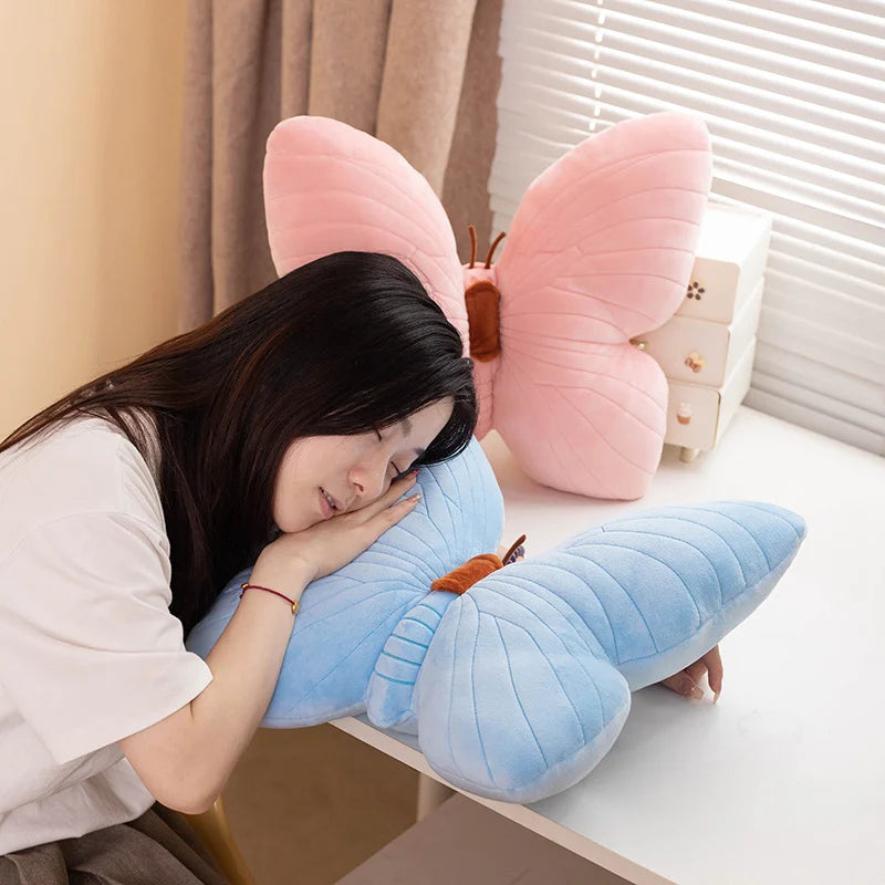 Person lying on a butterfly-shaped kawaii pillow with pink and blue wings in a room.