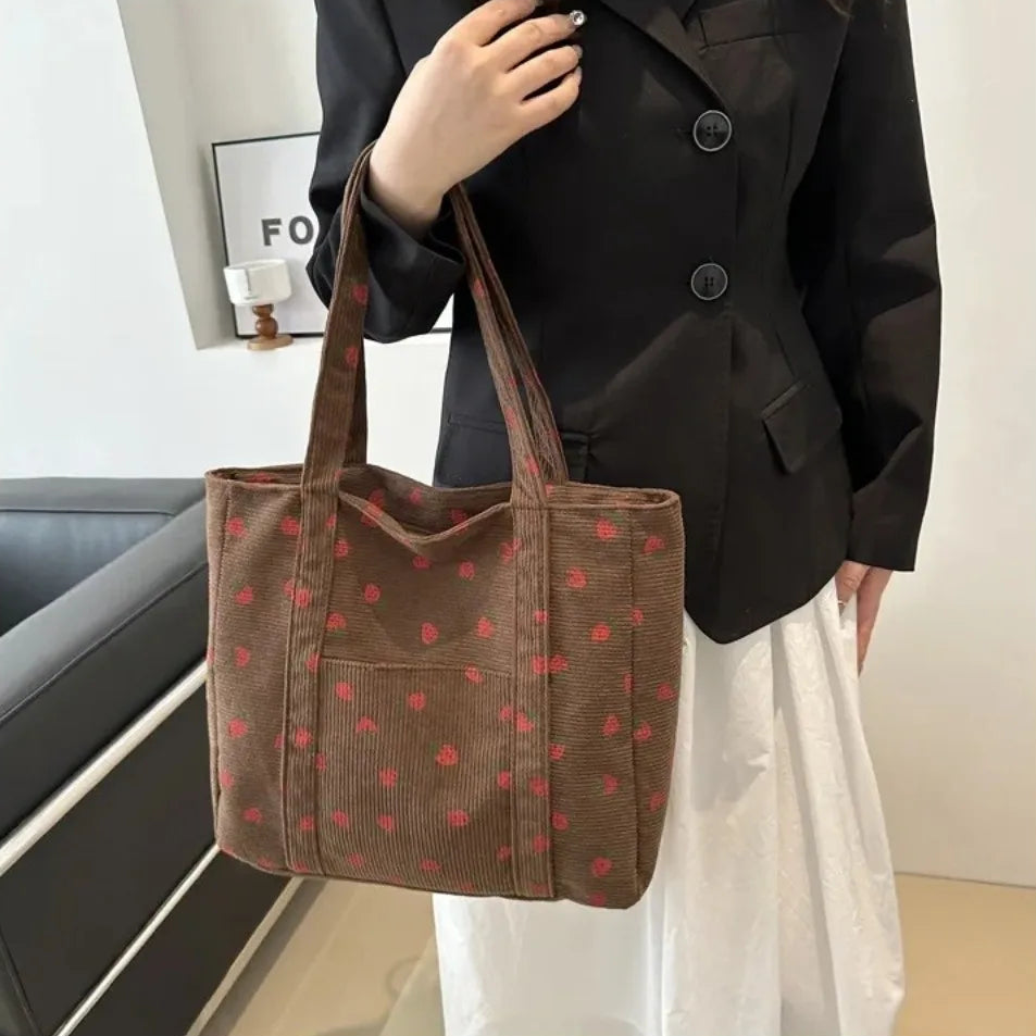 A woman wearing a black coat holds up a large brown tote bag with a red strawberry print in an indoor setting.