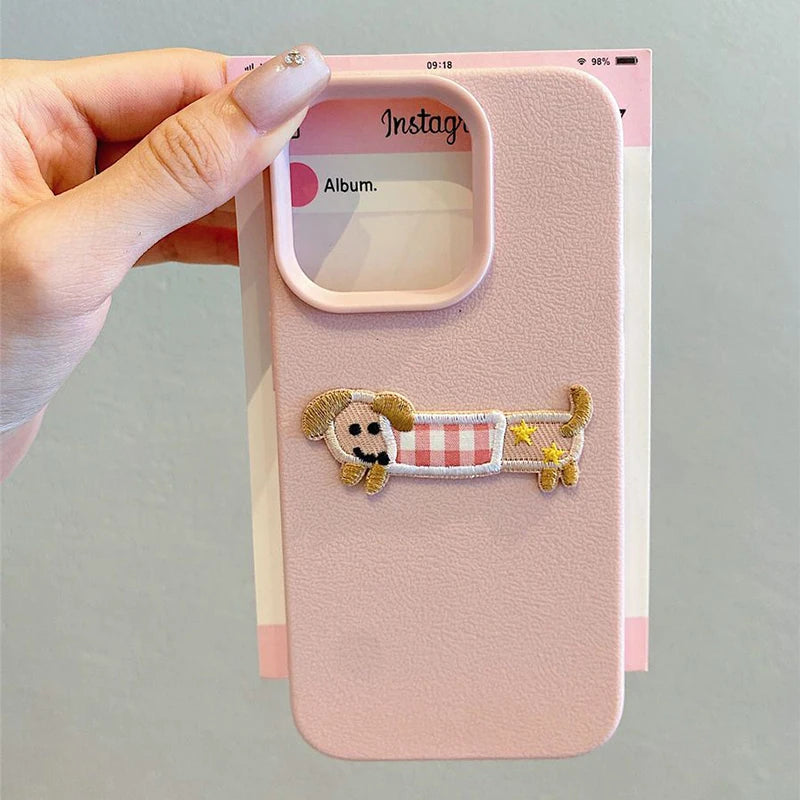 Pink phone case with a kawaii cartoon design of a dog on a checkered blanket, held by a hand.