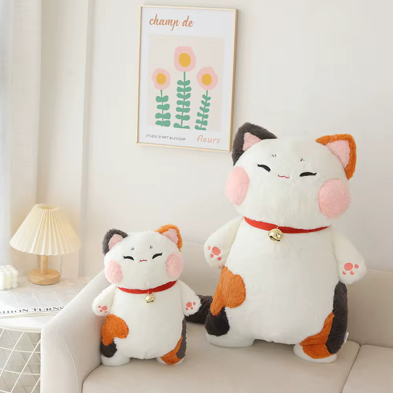 Two white cat plush toys with pink, orange, and black patches, one sitting on a beige sofa and one larger one standing beside it, in a softly lit room with a framed floral print on the wall.