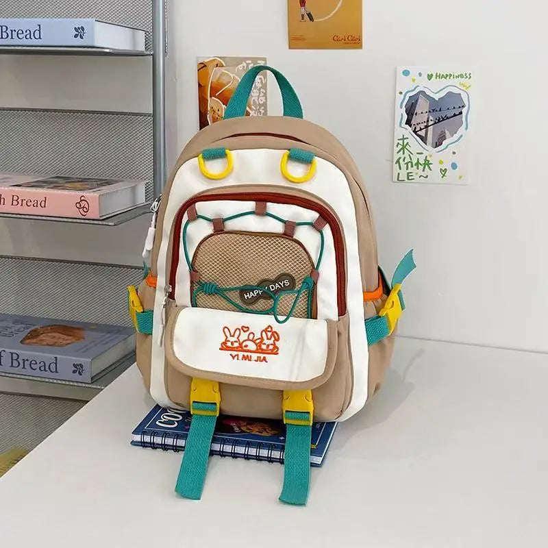 A beige and white backpack with teal straps and yellow accents, styled on a white table with books next to a shelf.