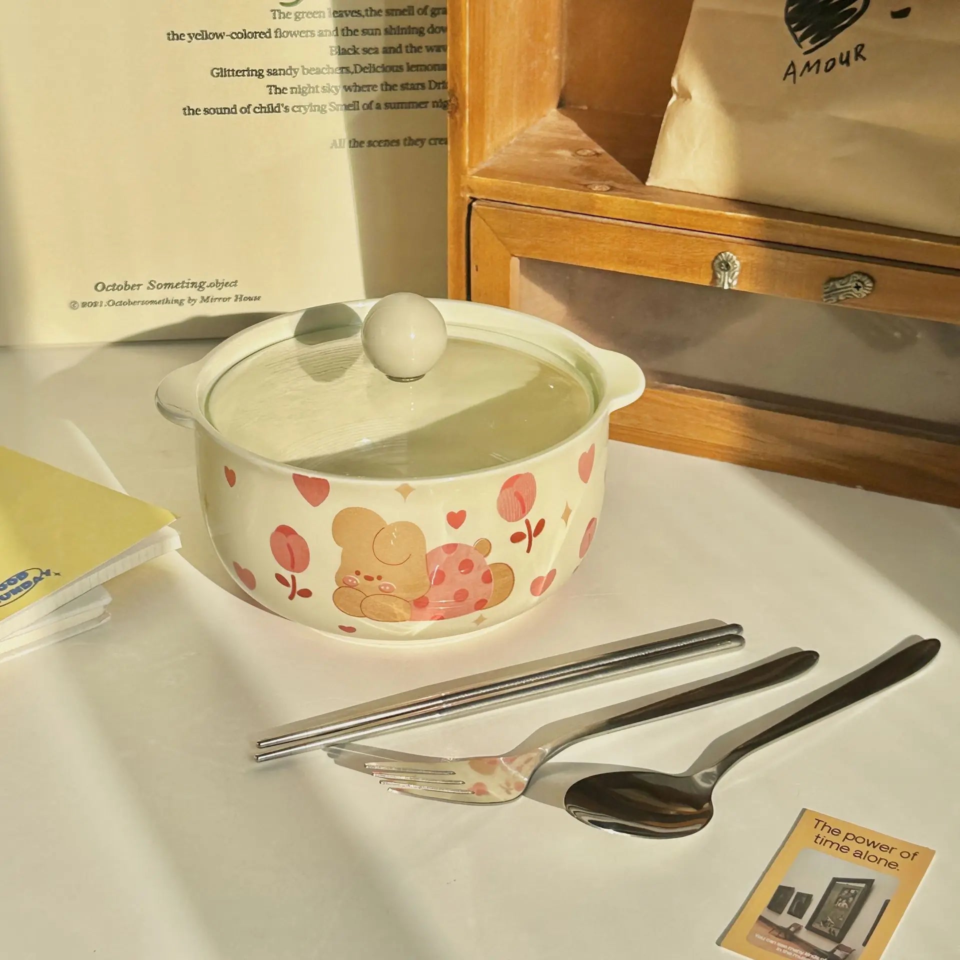 Ceramic kawaii ramen bowl with a lid, spoon, and fork on a table with books in the background