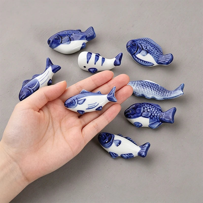 Hand holding a small blue and white fish kawaii chopstick holders with more fish figurines on a gray background