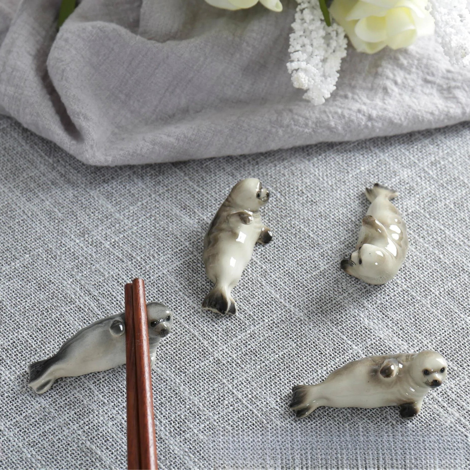 Four ceramic seal kawaii chopstick holders on a textured surface with a cloth and flowers in the background.