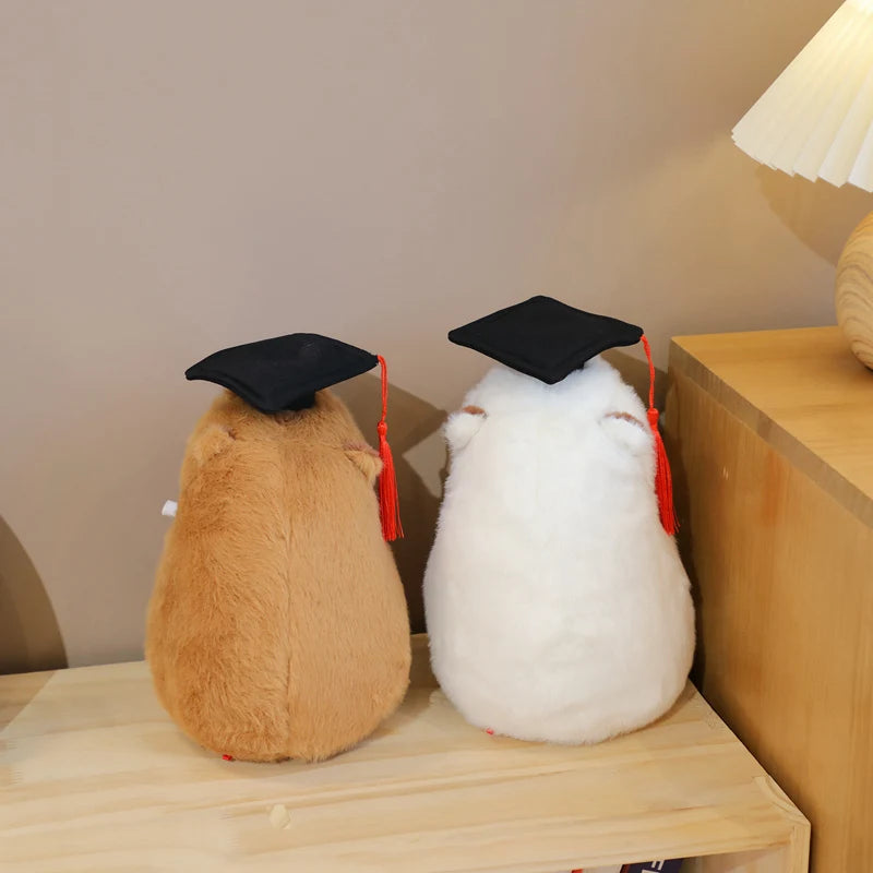 Two capybara plush toys, one brown and one white, wearing graduation caps on a wooden surface.