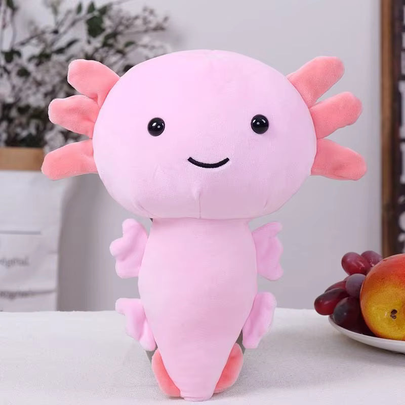 A pink axolotl plush toy with a cute stitched smiley face, black eyes, and small gills, standing on a white surface with a plate of fruit in the background.