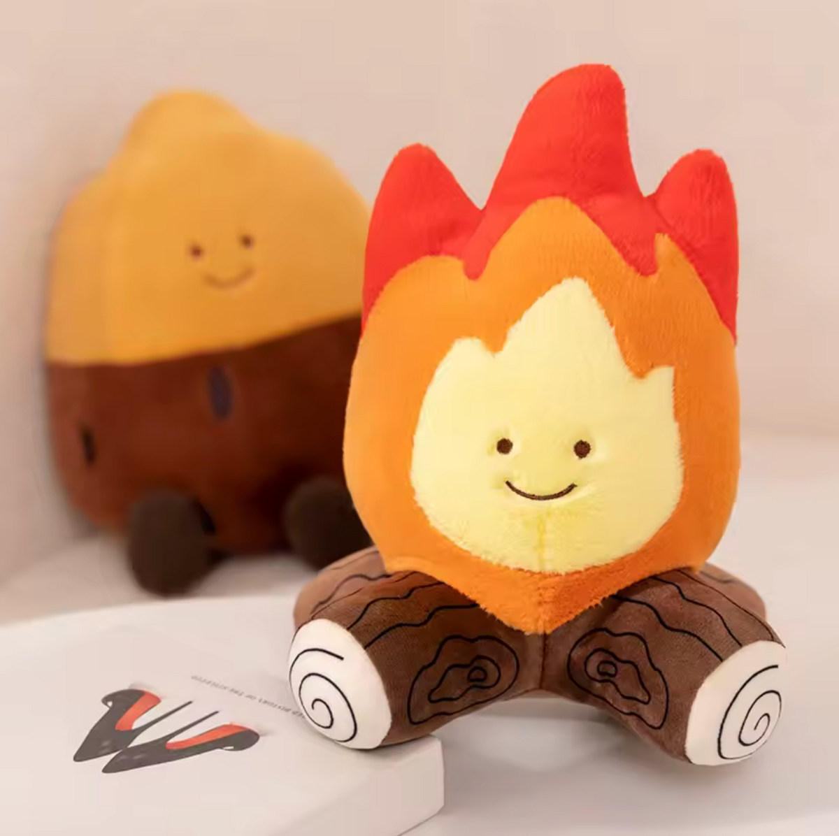 A cute flame-shaped plush toy sitting on logs, with a second similar plush toy blurred in the background. The toy has a yellow face and chest, red and orange flames on top, and brown logs with white spiral ends.