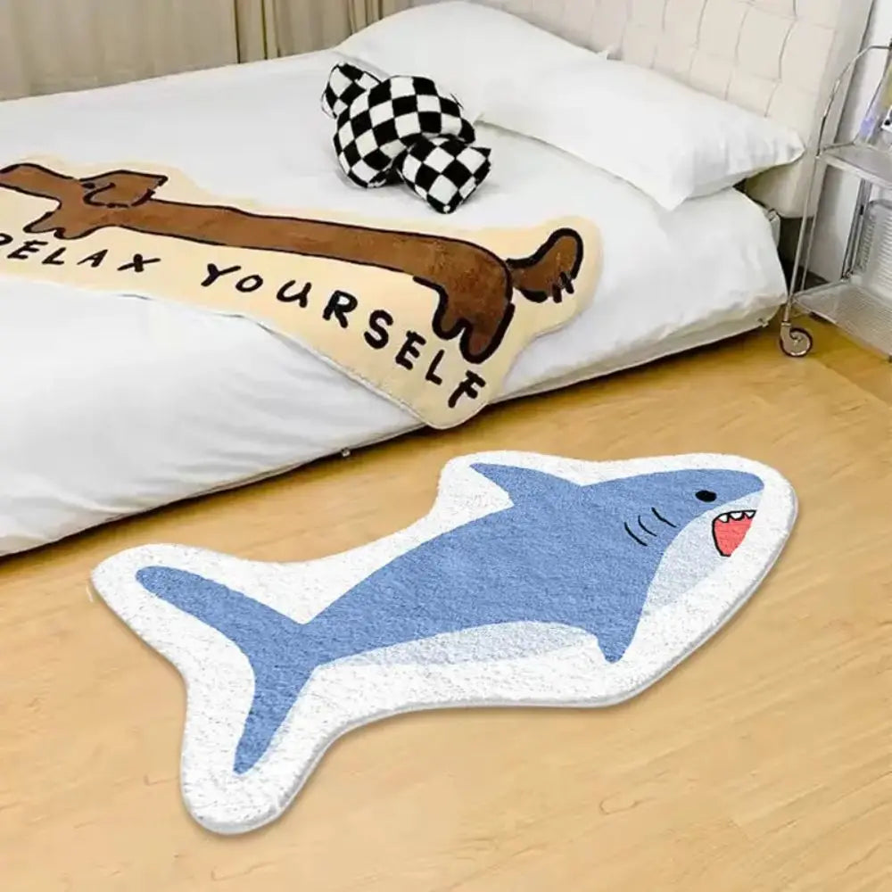 Shark-shaped kawaii rug on a wooden floor next to a bed with a blanket and pillow.