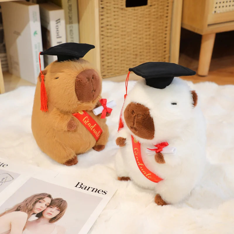 Two capybara plush toys resembling animals wearing graduation caps and gowns on a surface with a magazine underneath.