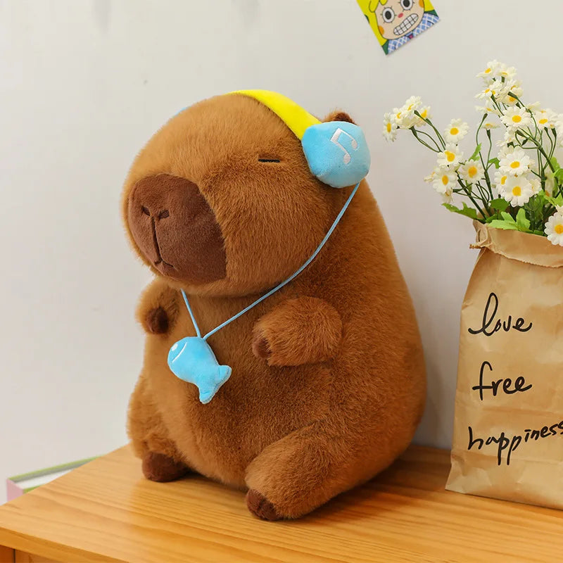 Capybara plush toy with headphones on a wooden surface next to a bag with flowers and text.