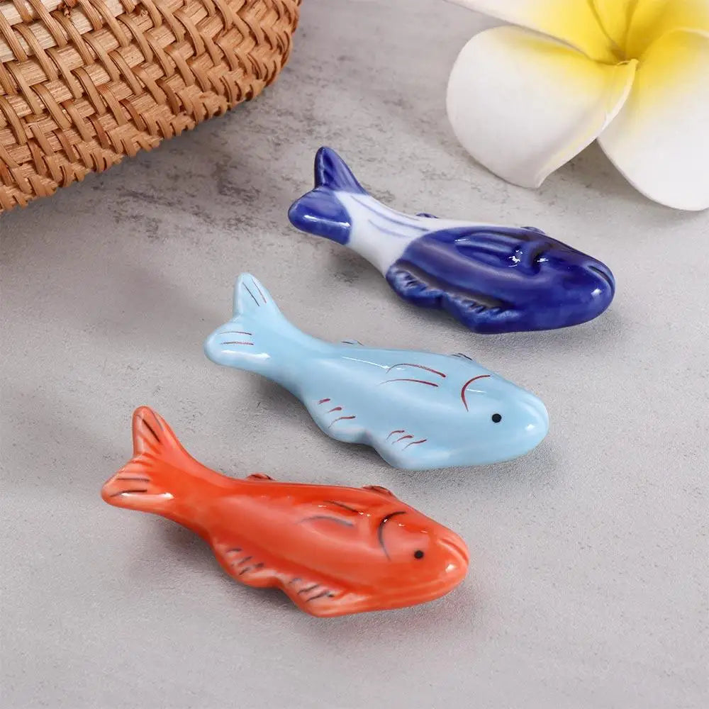 Three ceramic fish-shaped kawaii chopsticks holders in red, blue, and light blue on a gray surface with a woven basket and flower in the background.