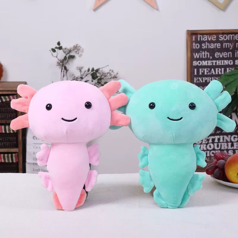 Two plush axolotl toys on a white tabletop in a styled indoor setting. One is light pink with darker pink gills and feet, and the other is mint green with darker green gills and feet.