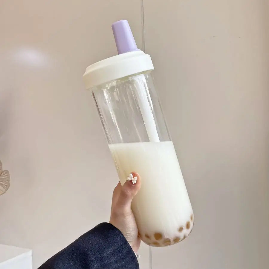 Hand holding a clear kawaii cup with a white lid filled with a creamy beverage, possibly bubble tea, against a neutral background.