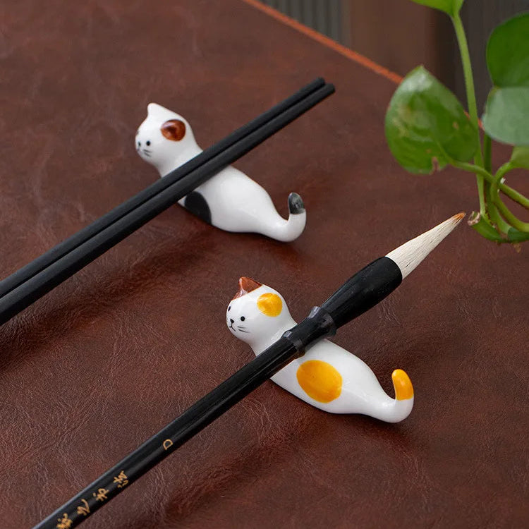 Two ceramic cat-shaped kawaii chopstick holders holders on a wooden surface with a plant in the corner.