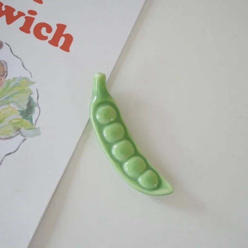 Green ceramic pea pod kawaii chopstick holderon a white surface with a children's book in the background.