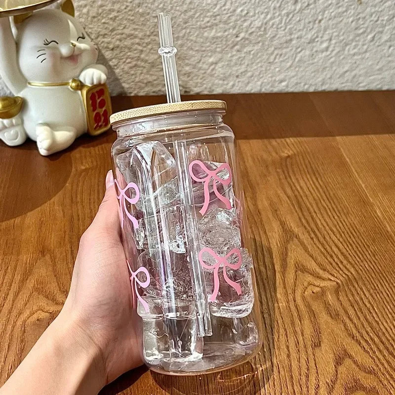 Clear glass kawaii cup with pink bows and a straw lid held by a hand on a wooden surface.