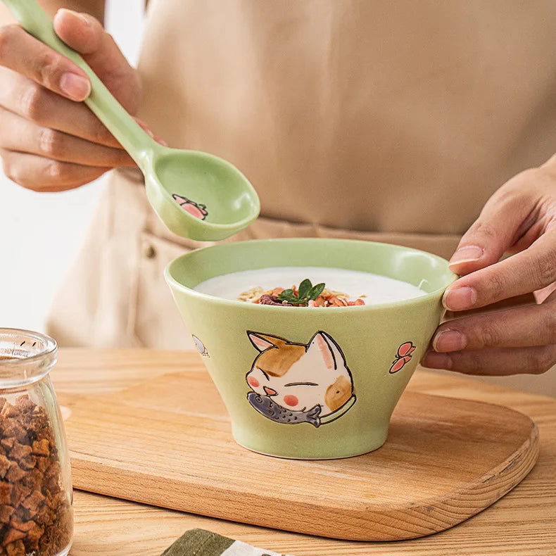 Green ceramic kawaii ramen bowl with cat design on a wooden surface, being filled with a white liquid.