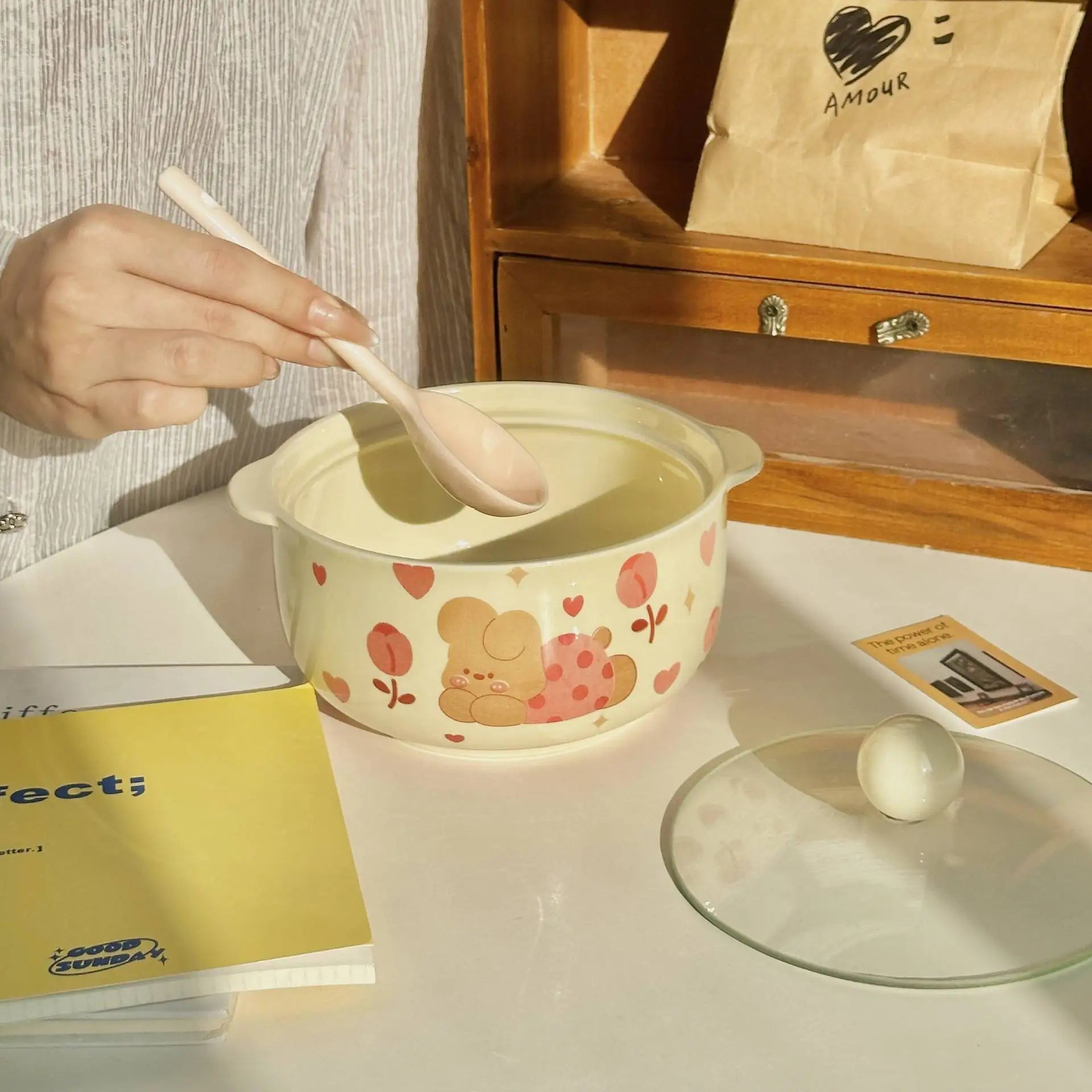 Person using a spoon to eat from a cartoon-themed kawaii ramen bowl on a table with a magazine and glass.