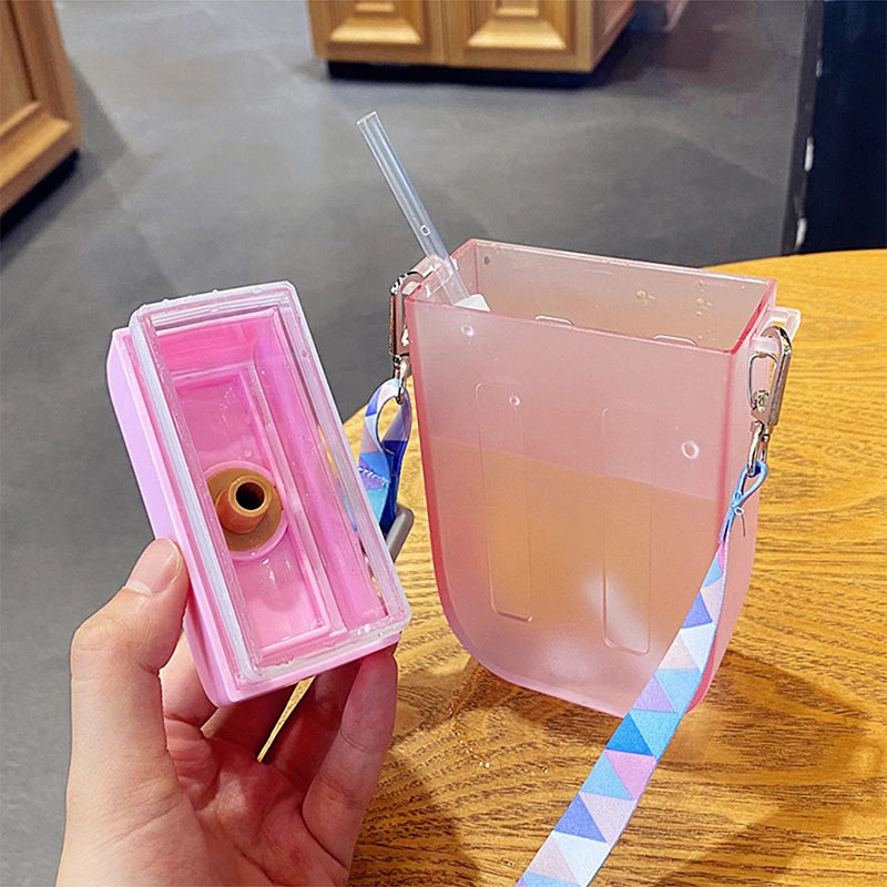 Pink transparent kawaii water bottle with a straw and colorful strap held by a hand on a wooden surface.