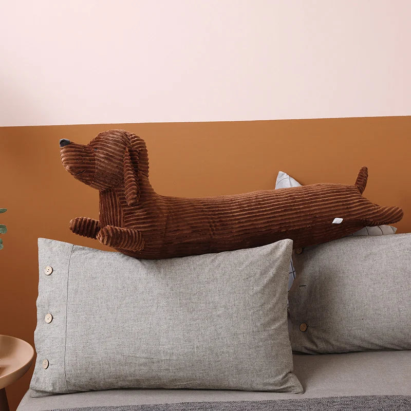 Brown dog-shaped kawaii pillow on a gray pillow against a brown wall.