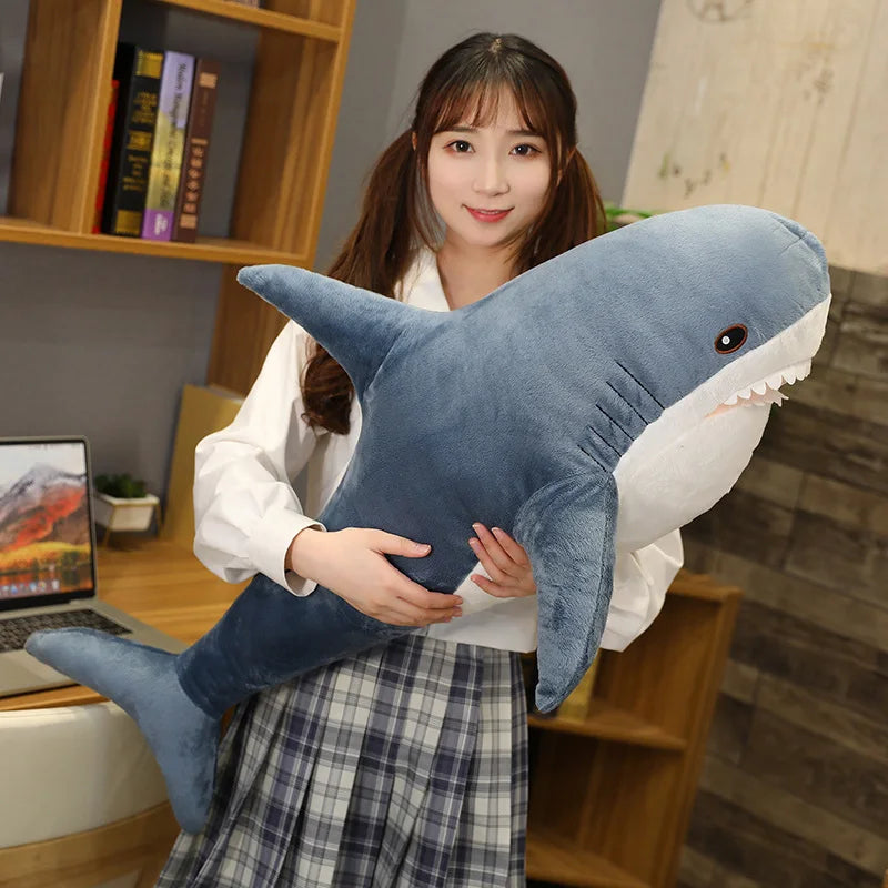Person holding a large shark-shaped kawaii plush toy in a room with a bookshelf and laptop.