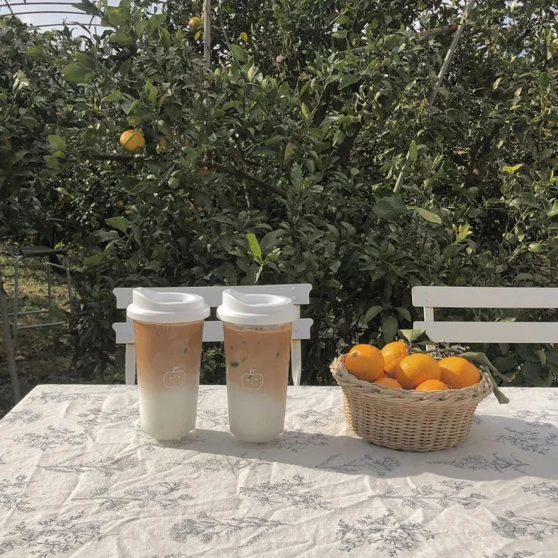 Two coffee Kawaii cups with a white lid on a table outdoors, with a basket of oranges and greenery in the background.