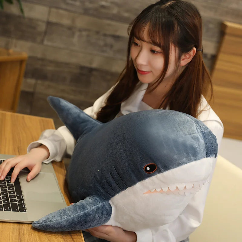 Person sitting at a table with a laptop, holding a large shark-shaped kawaii plush toy.