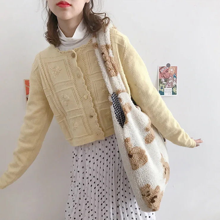 Person wearing a beige kawaii cardigan and holding a patterned bag against a plain background