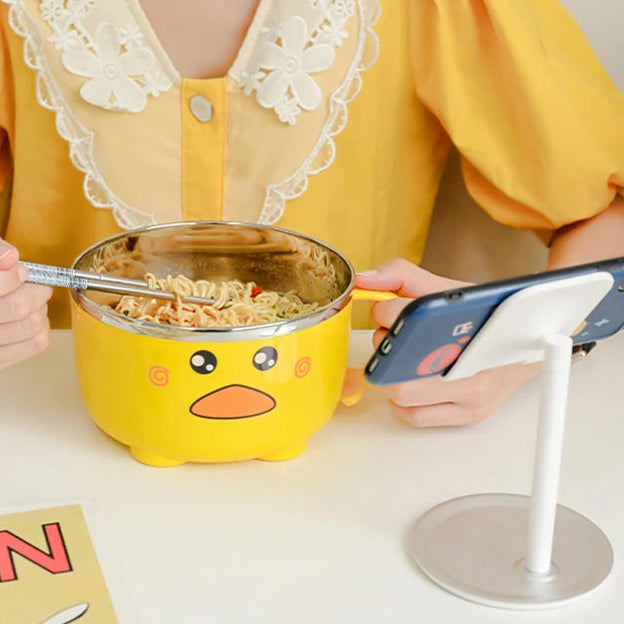 Person in yellow outfit eating from a yellow cartoon-style kawaii ramen bowl with a phone on a stand.