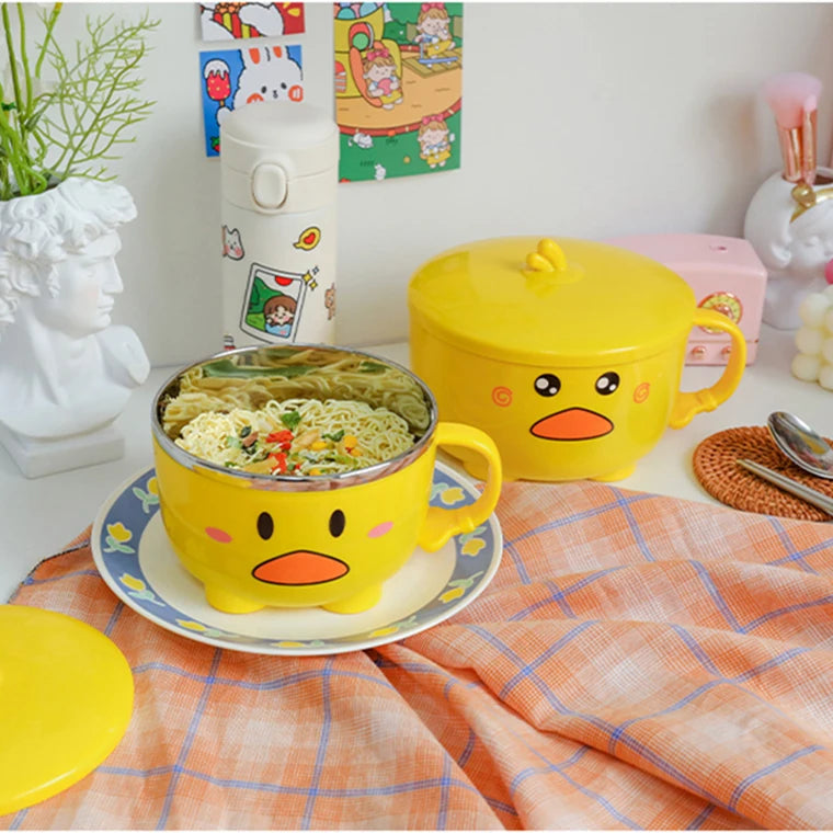 Yellow duck-shaped kawaii ramen bowls with a lid on a checkered tablecloth, surrounded by decorative items.