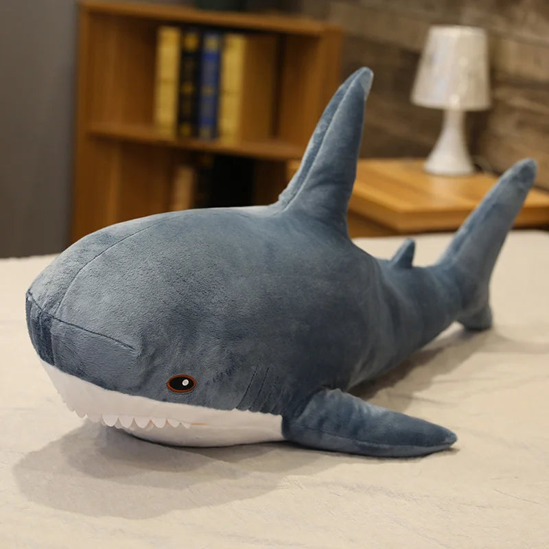 Large kawaii plush shark toy on a bed with a bookshelf and lamp in the background