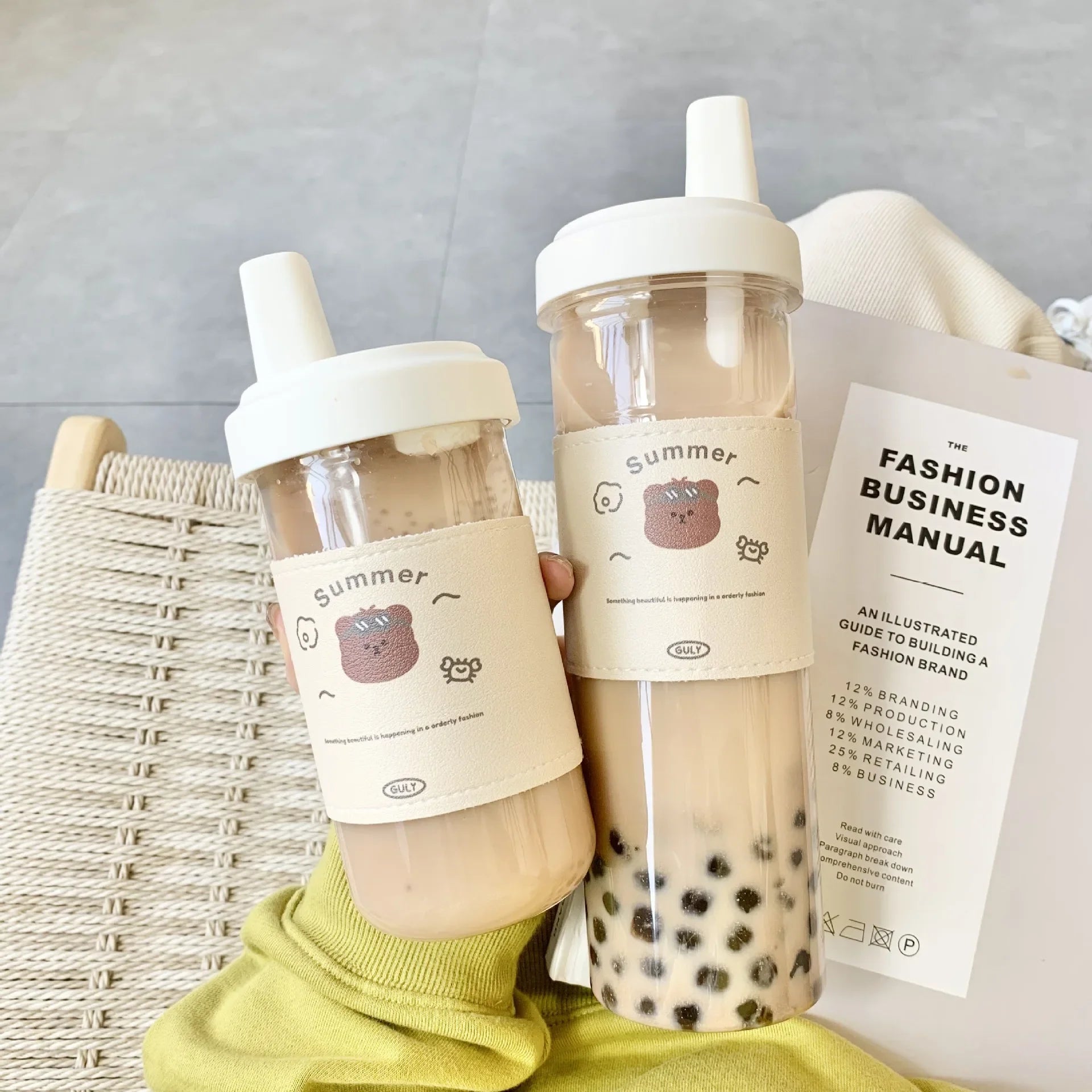 Two bubble tea kawaii cups with cute labels and a book titled 'The Fashion Business Manual' on a light gray background.