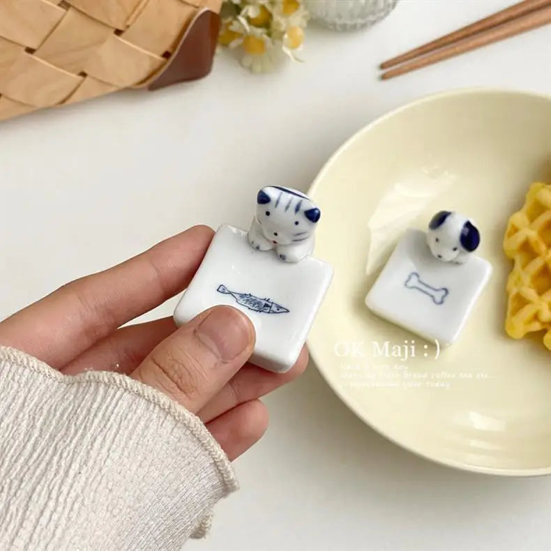 Hand holding a small white kawaii chopstick holders with a cat design, next to a plate with similar containers.