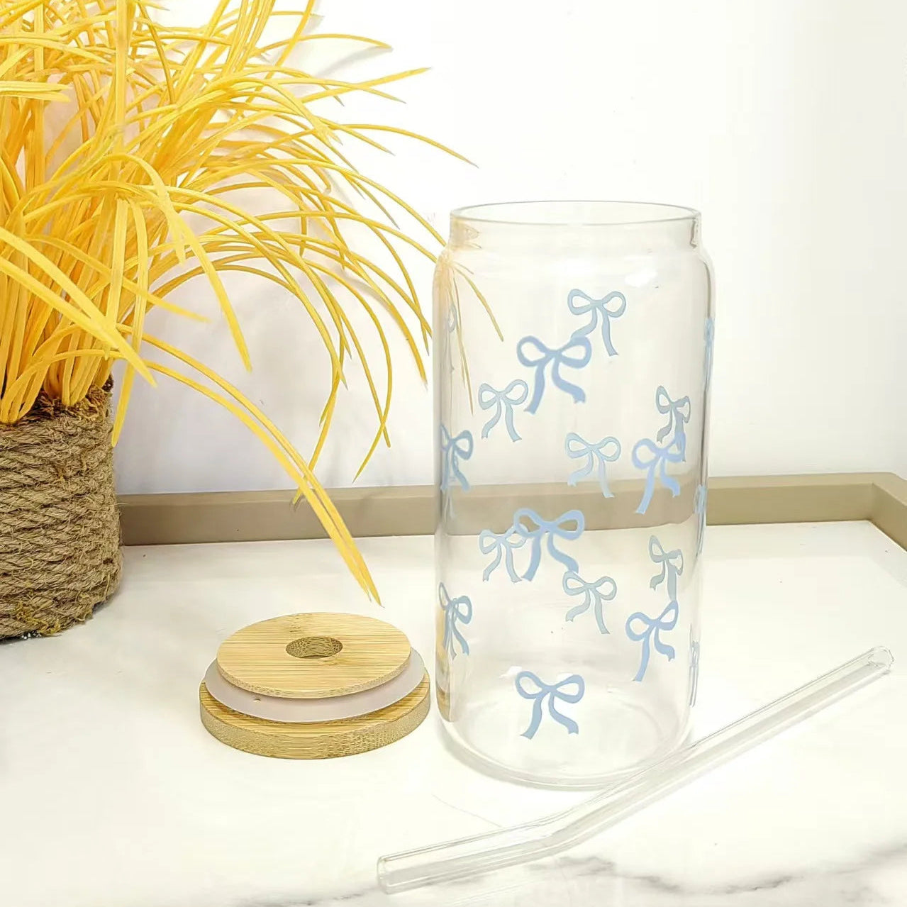 Clear glass kawaii tumbler with blue bow designs, wooden lid, and straw on a white surface with yellow plant.