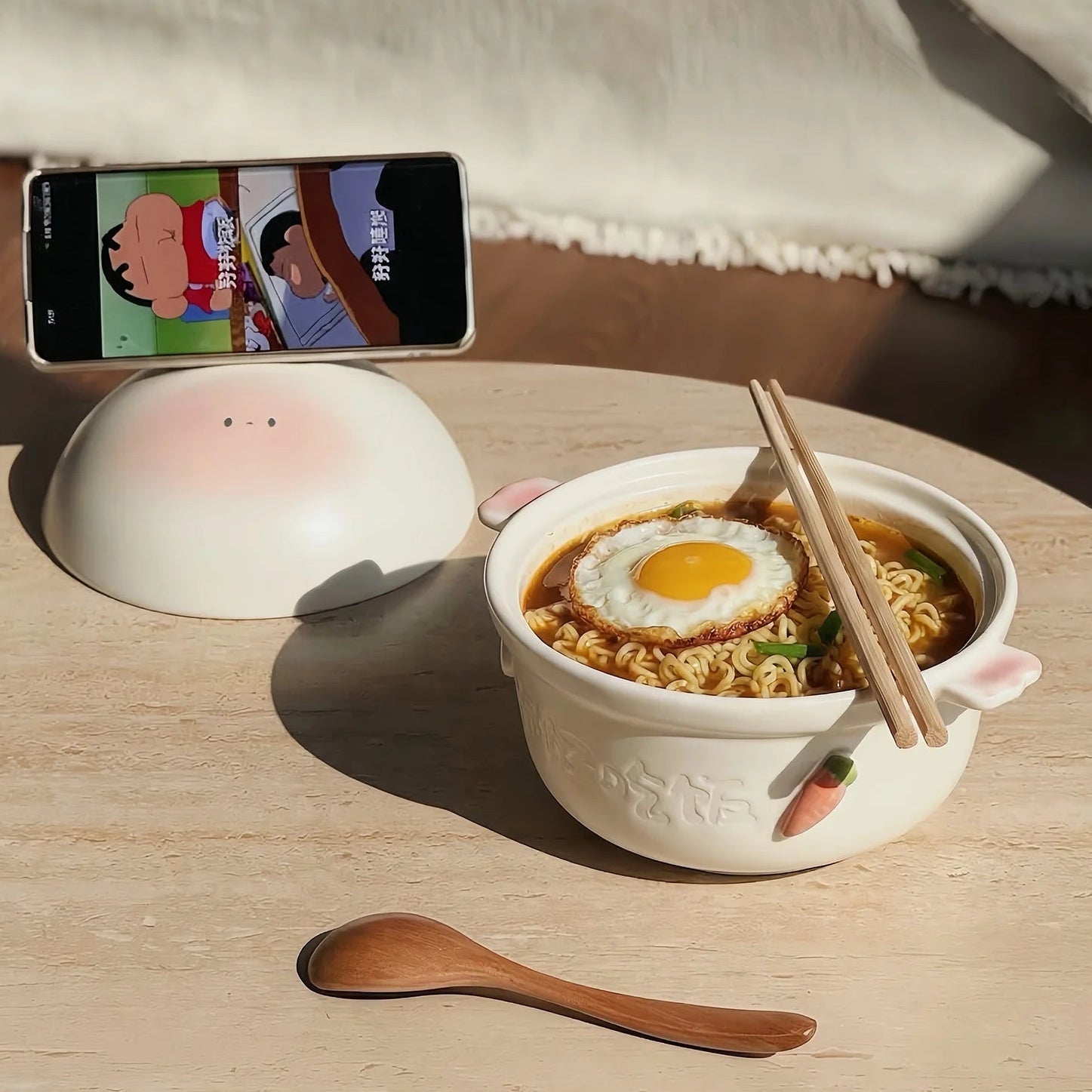 Noodle kawaii bowls with an egg and chopsticks on a table next to a smartphone displaying a cartoon.