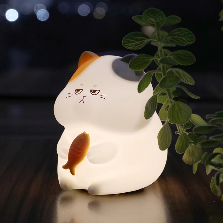 Cat-shaped kawaii night light with a fish figure, illuminated in a dark setting with a plant.