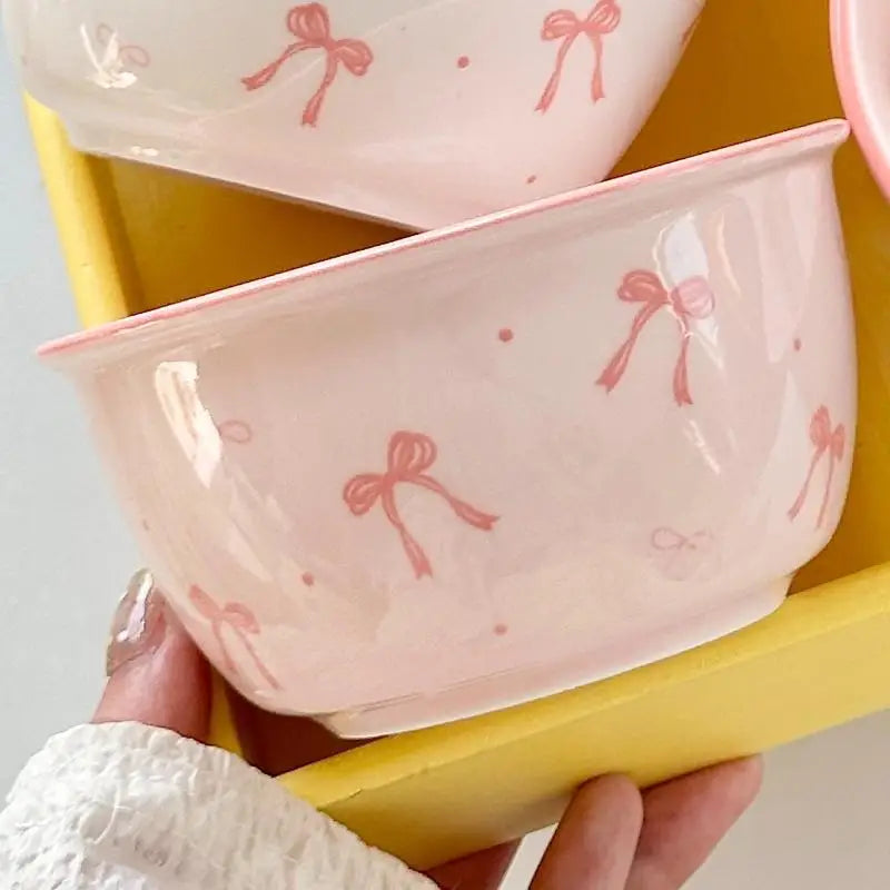 A close-up of a hand holding a light pink ceramic bowl decorated with a repeating red bow pattern, with other matching bowls stacked in a yellow container behind it.