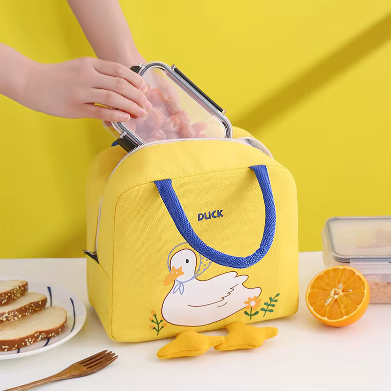 A yellow insulated lunch bag with blue handles and a duck illustration on the front, shown with a person's hands opening the lid and food items around it on a white table against a bright yellow background.