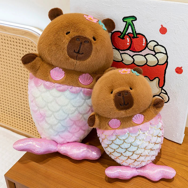 Two capybara plush toys with mermaid tails on a wooden surface.