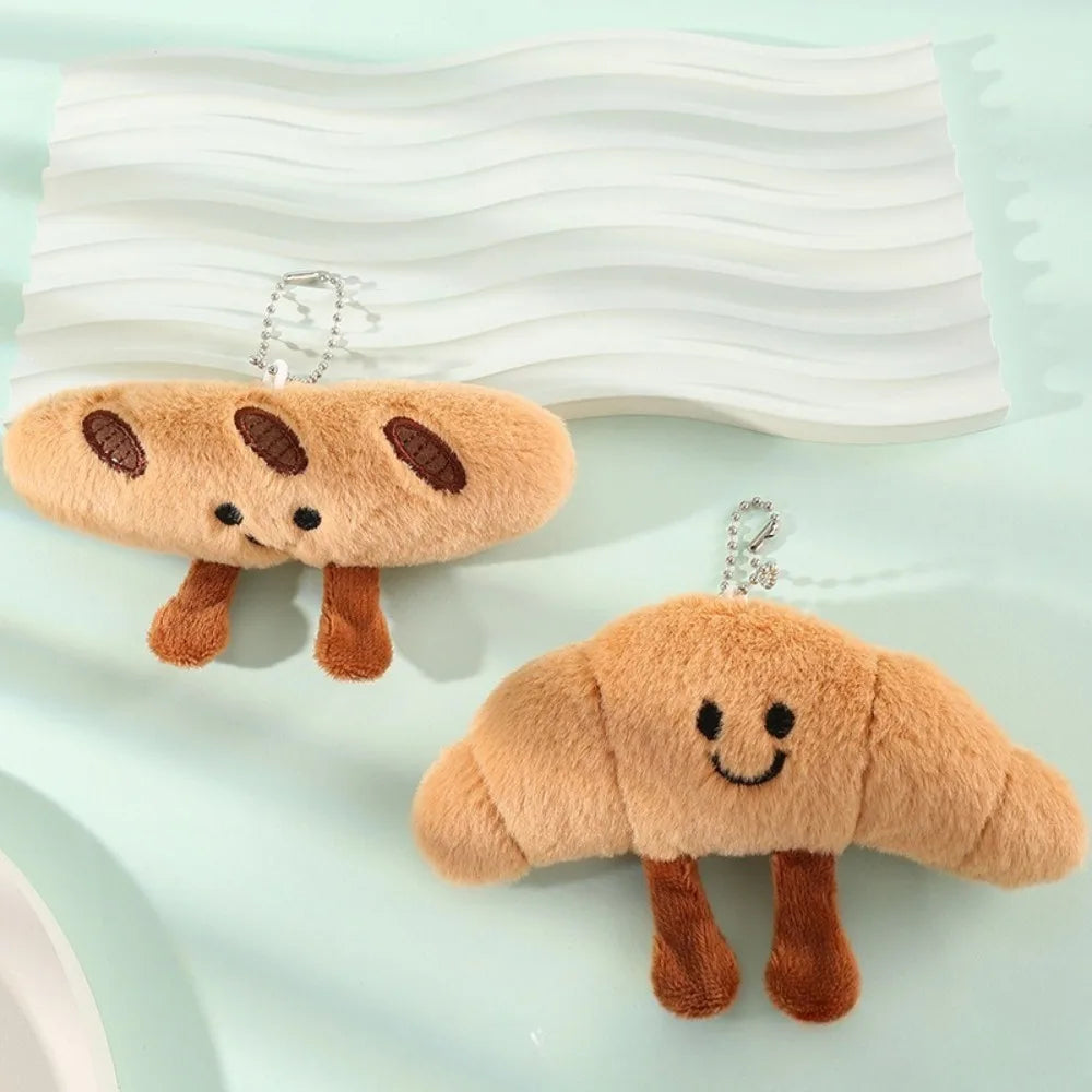 Two plush croissant-shaped keychain charms with embroidered smiling faces and brown legs, displayed on a pale mint background with a white wavy tray behind them.